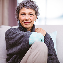 An elderly woman wearing a grey cable knit sweater and khakis, clutches her shoulder and smiles while seated on a white couch.