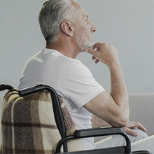 An elderly caucasian man is seated in a wheelchair and staring off to the right while in the thinker's pose.