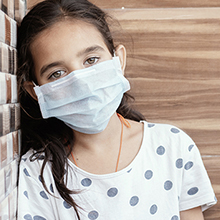 A young girl looks into the camera while wearing a white surgical mask